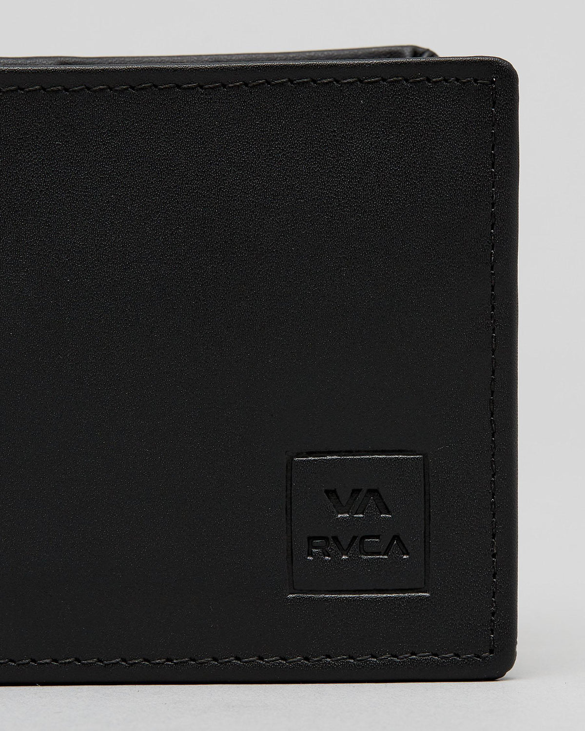 Close-up of a black leather bi-fold wallet showing the corner, black stitching, and an embossed square logo patch with 'VA RVCA' on a light gray background. cedar bifold