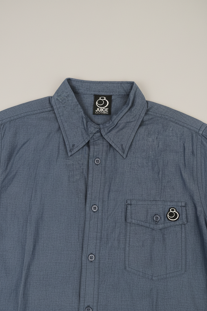 A close-up of a blue-gray button-up shirt with a pointed collar, a chest pocket, and a small circular metal ring detail on the pocket flap. The inside collar shows a black brand label reading 'JUDGE'.