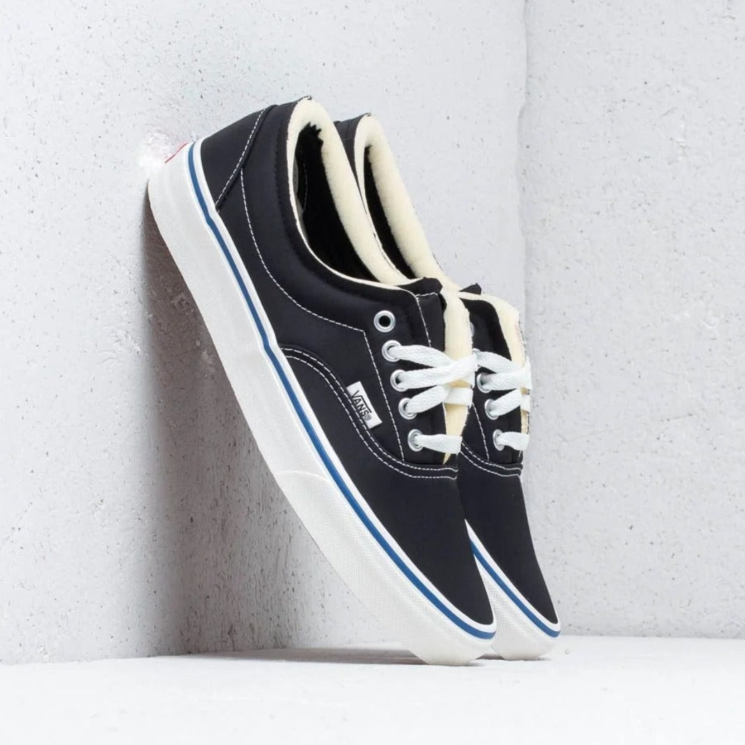 A pair of black Vans platform sneakers with white laces, white rubber soles, and a thin blue stripe, leaning against a light gray concrete wall.