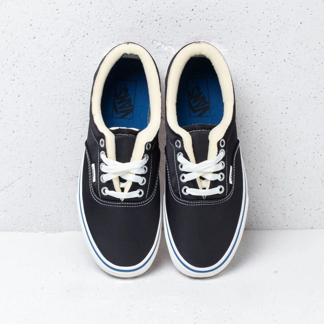 A pair of Vans sneakers in black canvas with cream laces, cream collar lining, and a blue Vans logo printed on the tongue insoles, set against a light gray textured background.