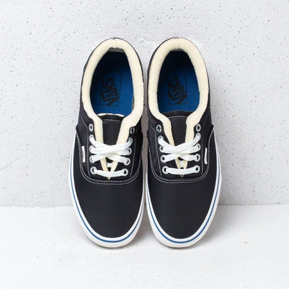 A pair of Vans sneakers in black canvas with cream laces, cream collar lining, and a blue Vans logo printed on the tongue insoles, set against a light gray textured background.