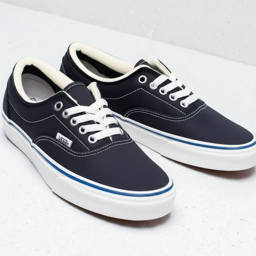 A pair of Vans Era low-top sneakers in navy canvas with white laces, white rubber soles, and a thin blue stripe detail, displayed on a light gray textured surface.