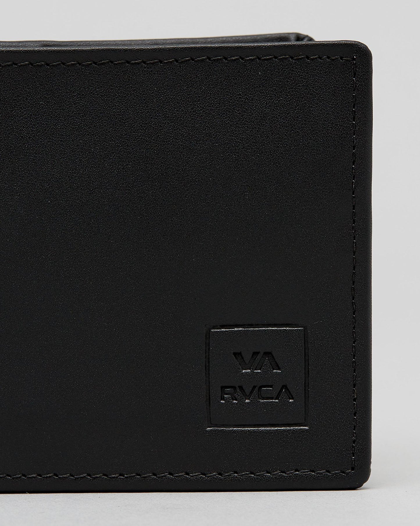 Close-up of a black leather bi-fold wallet showing the corner, black stitching, and an embossed square logo patch with 'VA RVCA' on a light gray background. cedar bifold
