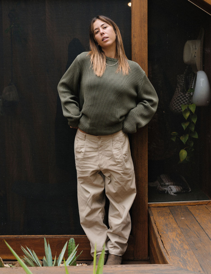 Woman wearing a green sweater and beige pants standing in front of a dark wall.