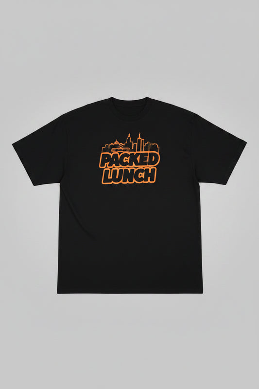 A black short-sleeve t-shirt hanging on a hanger against a plain off-white wall. The back of the shirt features a graphic print with an orange outline of a city skyline and the words 'PACKED LUNCH' in bold orange letters.