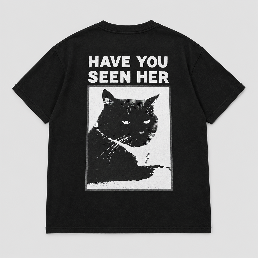Back view of a black short-sleeve T-shirt laid flat, featuring a large graphic print of a black cat inside a white rectangular frame with the text 'HAVE YOU SEEN HER' in white above it, on a light gray background.