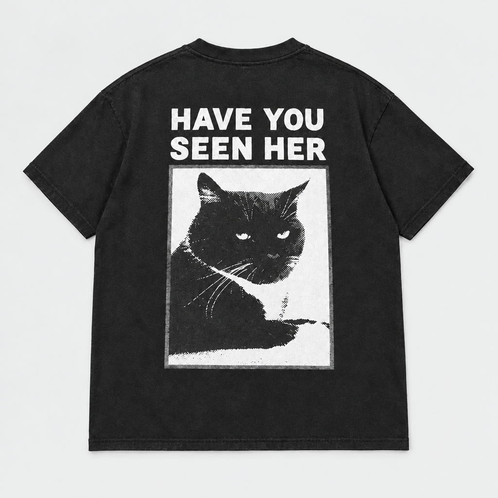 Back view of a black short-sleeve T-shirt laid flat, featuring a large graphic print of a black cat inside a white rectangular frame with the text 'HAVE YOU SEEN HER' in white above it, on a light gray background.