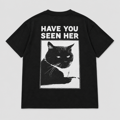 Back view of a black short-sleeve T-shirt laid flat, featuring a large graphic print of a black cat inside a white rectangular frame with the text 'HAVE YOU SEEN HER' in white above it, on a light gray background.