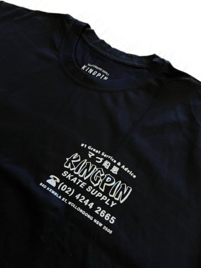 A close-up of a black t-shirt featuring white printed graphics and text on the front, including 'KINGPIN SKATE SUPPLY' and Japanese characters, with the brand 'KINGPIN' visible inside the collar.