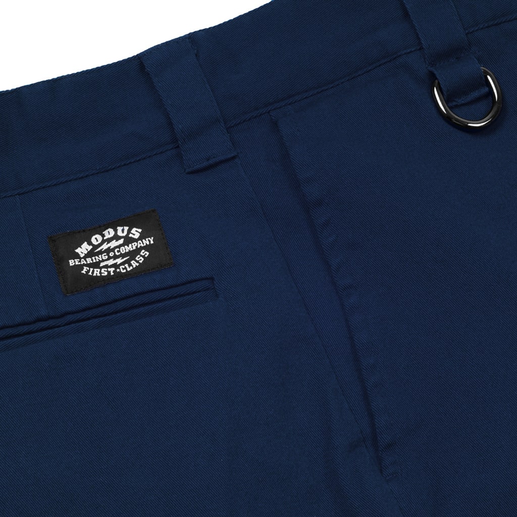 Close-up of navy blue trousers showing the waistband, belt loop, pocket, and a black woven brand patch that reads 'MOCUS BEARINGS COMPANY FIRST CLASS'.
