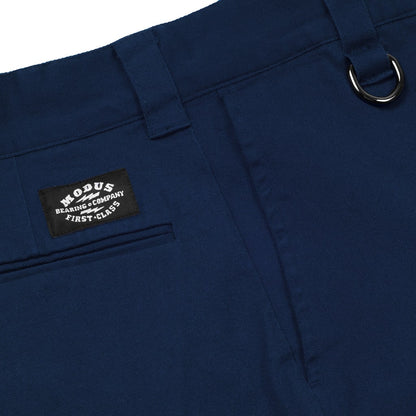 Close-up of navy blue trousers showing the waistband, belt loop, pocket, and a black woven brand patch that reads 'MOCUS BEARINGS COMPANY FIRST CLASS'.