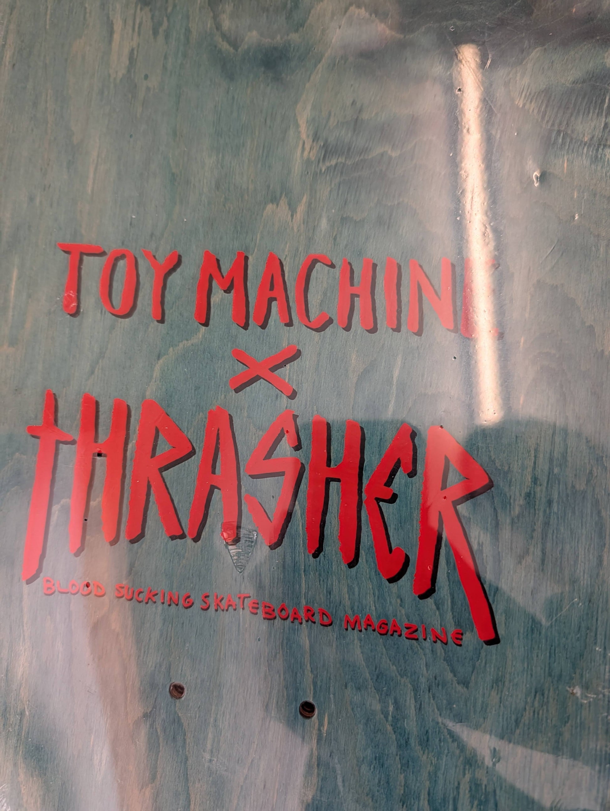 A close-up of a metallic teal-blue surface with bold red text reading 'TOY MACHINE x THRASHER' and smaller text 'BLOOD SUCKING SKATEBOARD MAGAZINE' underneath.