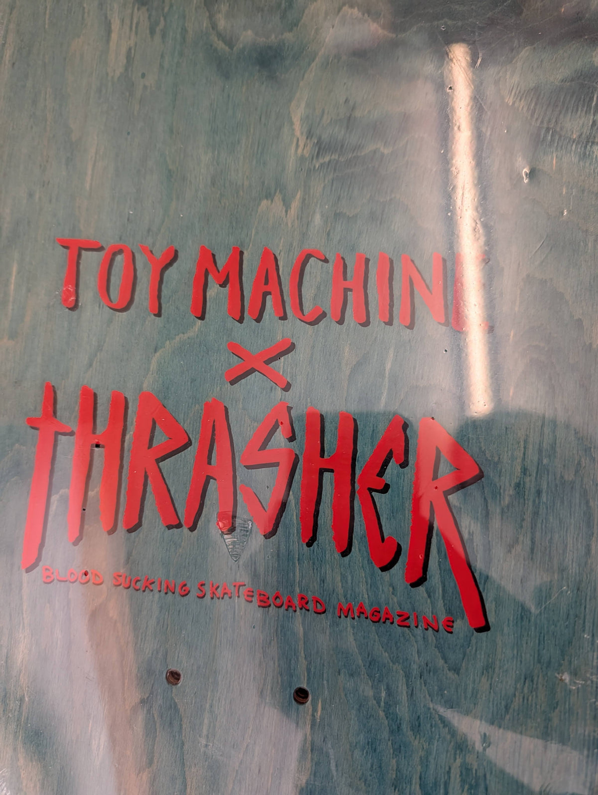 A close-up of a metallic teal-blue surface with bold red text reading 'TOY MACHINE x THRASHER' and smaller text 'BLOOD SUCKING SKATEBOARD MAGAZINE' underneath.