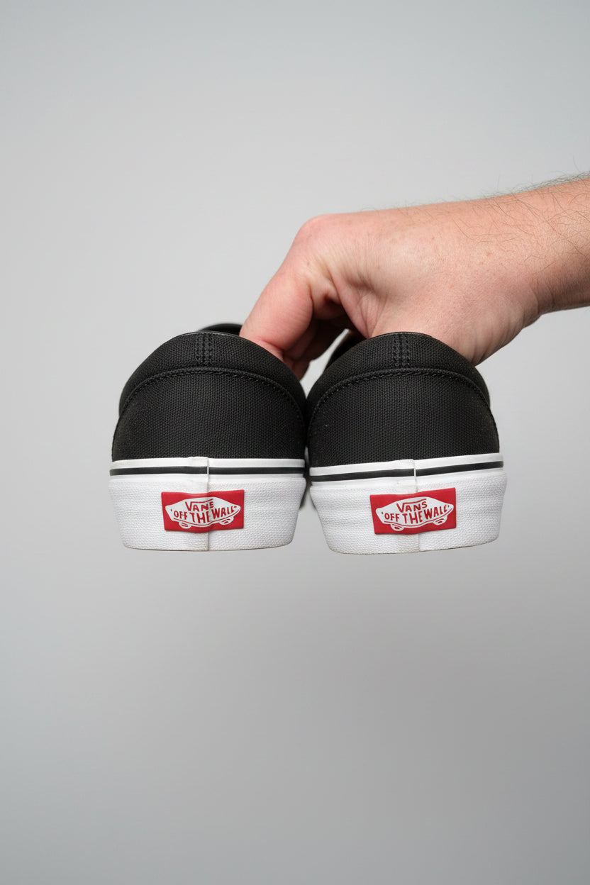 A close-up of a person's hand holding a pair of dark gray Vans Off-The-Wall slip-on sneakers against a concrete floor background.