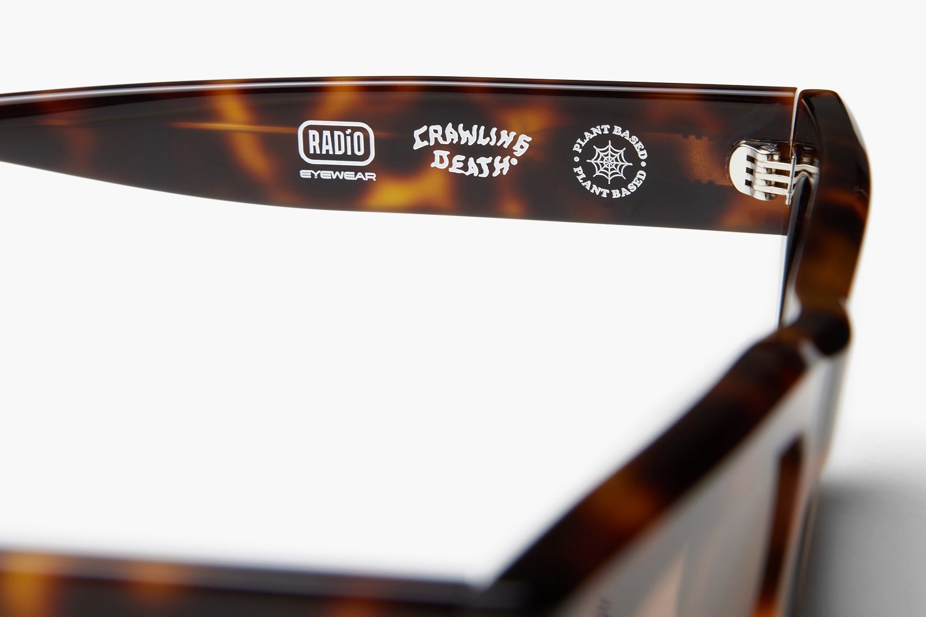 Close-up of the inside of a tortoiseshell eyeglass arm showing printed branding and logos, including RADIO EYEWEAR and Crowding Death, on a white background.