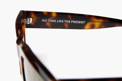 Close-up of a tortoiseshell eyeglass frame arm with the text 'NO TIME LIKE THE PRESENT' printed in white on a dark brown and amber patterned temple, against a plain white background.