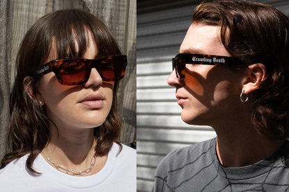 Two models wearing black-framed sunglasses with orange-tinted lenses. The left model has brown hair and is wearing a white top against a wooden wall, while the right model has dark hair and is wearing a gray shirt against a corrugated metal wall.