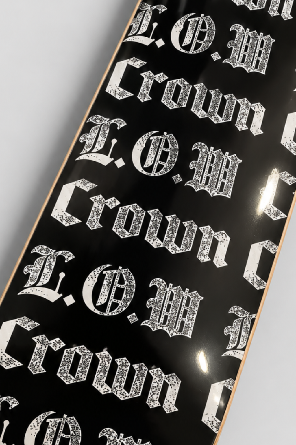 A skateboard deck with a glossy black background and a repeating white Gothic-style 'Crown' logo pattern, photographed from an overhead angle on a light gray background.