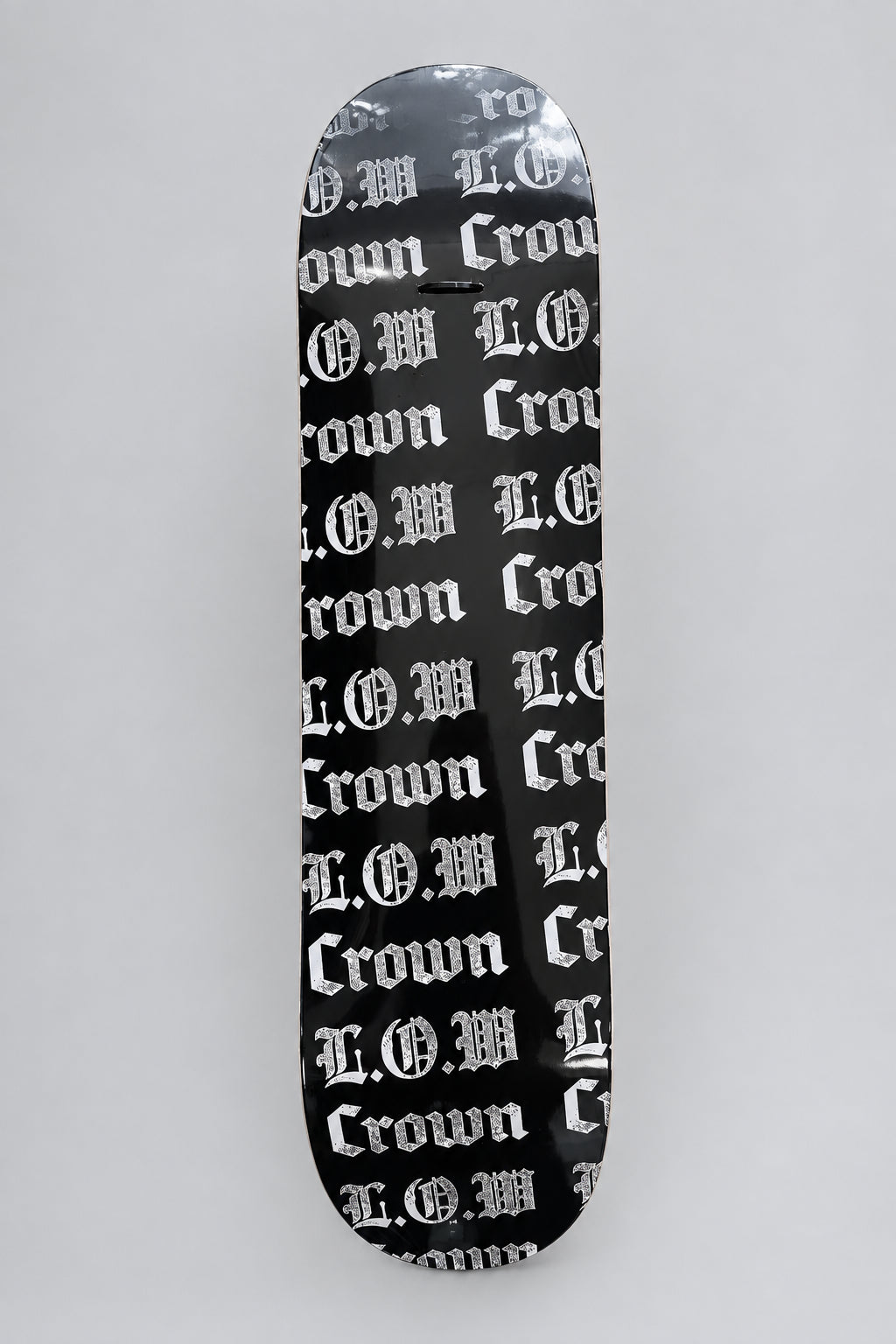 A skateboard deck with a black background and a repeating white Gothic 'Crown' and L.O.W logos repeated pattern, photographed from directly above on a light brown wood floor.