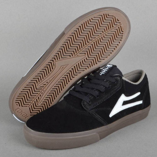 A pair of black suede skate shoes with a white side logo, shown with one shoe turned to display the brown treaded outsole against a neutral gray background.