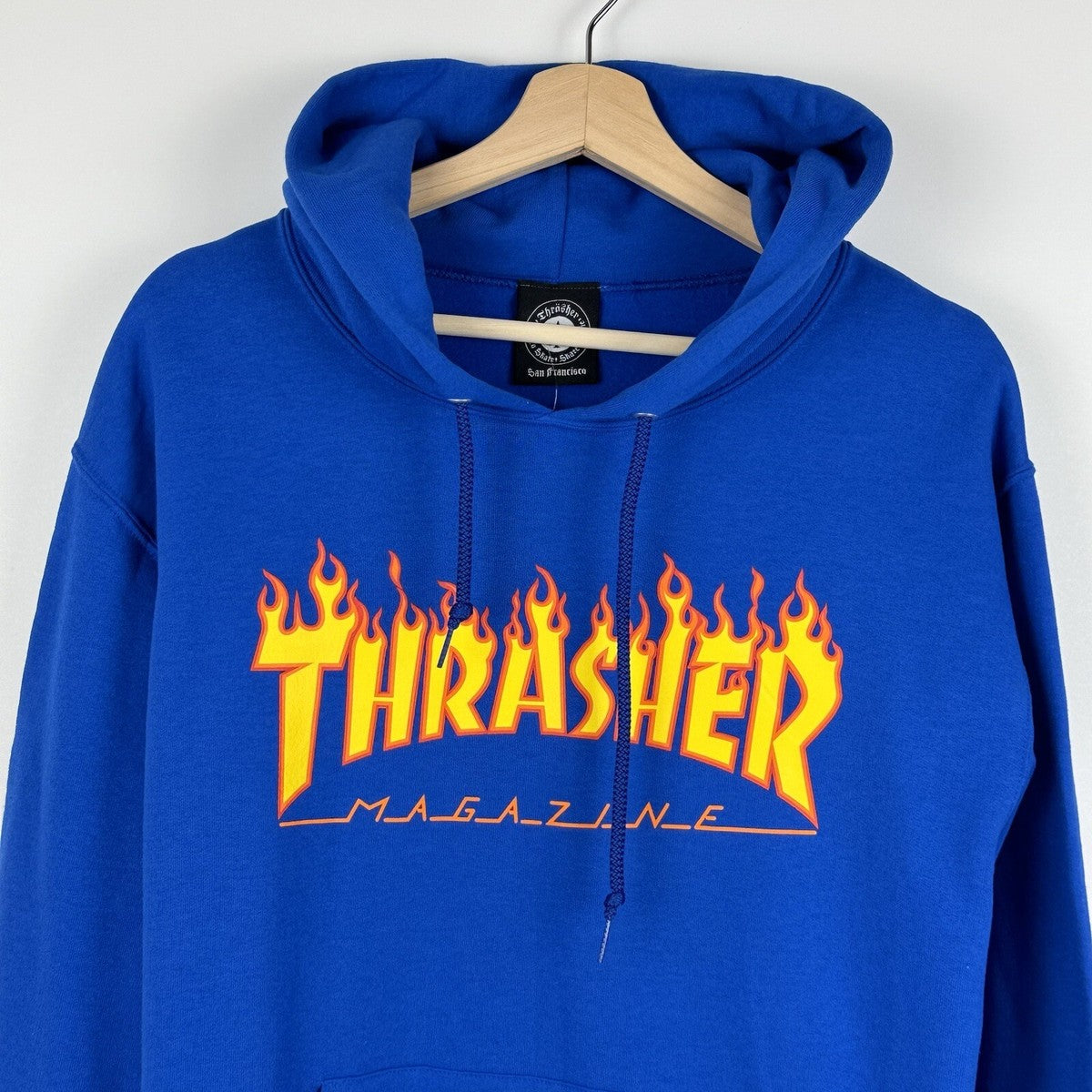 A royal blue Thrasher Magazine hoodie hanging on a wooden hanger against a light gray wall. The hoodie features the iconic yellow and orange flame 'THRASHER' logo with 'MAGAZINE' printed underneath.