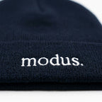 A dark navy blue knit beanie with the word 'modus.' embroidered in white on the cuff, resting on a light wood surface.