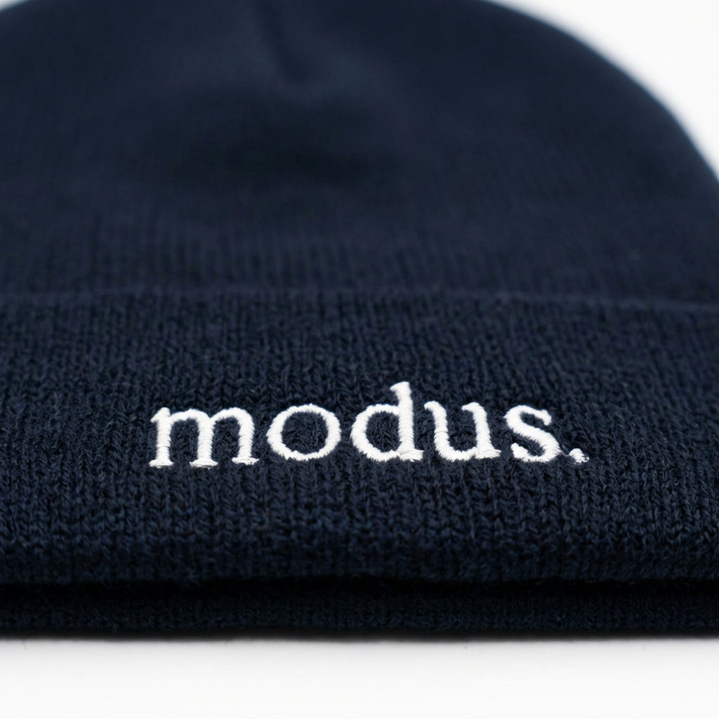 A dark navy blue knit beanie with the word 'modus.' embroidered in white on the cuff, resting on a light wood surface.