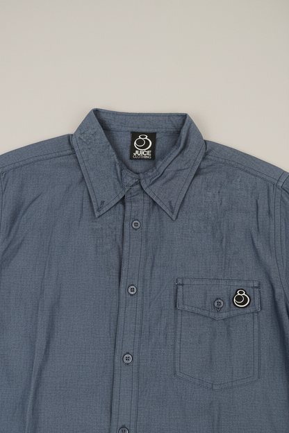 A close-up of a blue-gray button-up shirt with a pointed collar, a chest pocket, and a small circular metal ring detail on the pocket flap. The inside collar shows a black brand label reading 'JUDGE'.