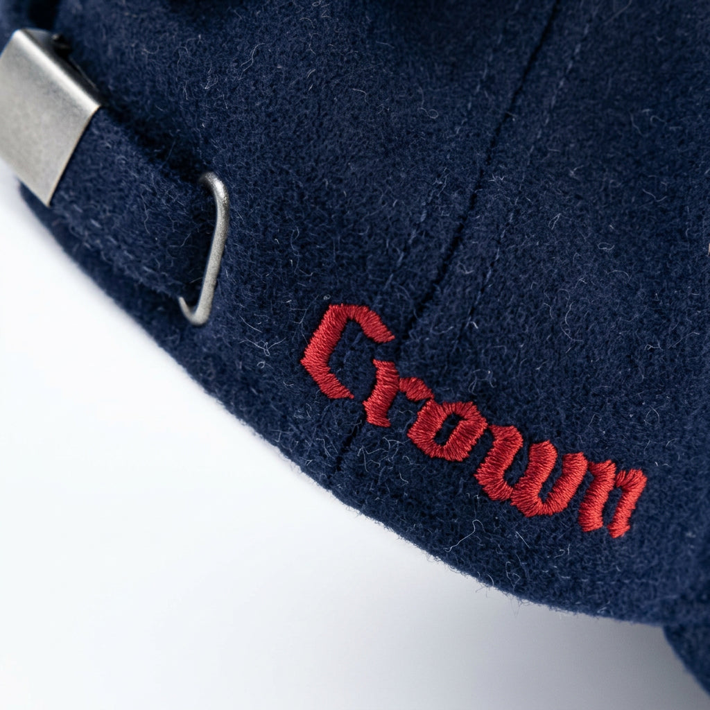 A close-up of a dark navy blue felt cap resting on a light wood surface, featuring red embroidered 'Crown' lettering and a silver metal buckle strap.