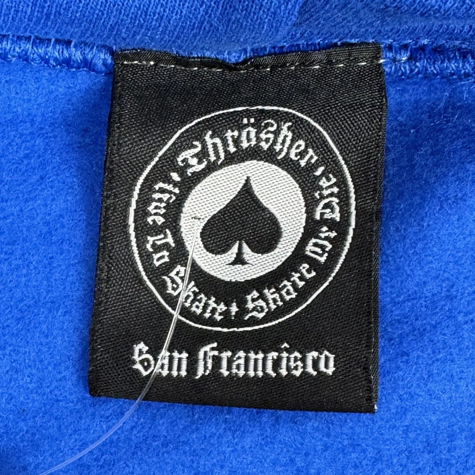 A close-up of a black fabric brand label sewn onto a bright blue garment. The label features a circular white graphic with a spade symbol.
