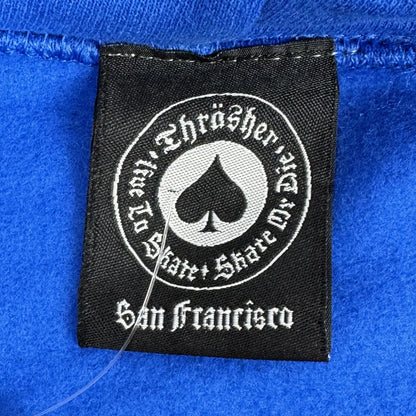 A close-up of a black fabric brand label sewn onto a bright blue garment. The label features a circular white graphic with a spade symbol.