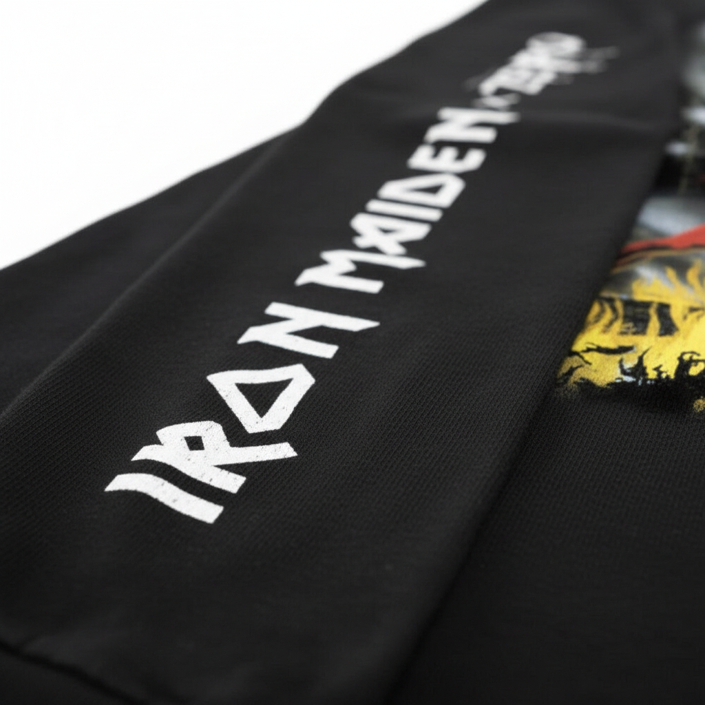 Close-up of a black garment featuring large white 'IRON MAIDEN' text printed diagonally, with a small yellow graphic visible on the right side.