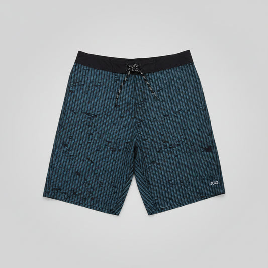 A pair of men's board shorts with a blue and black pixelated pattern, laid flat on a grey floor. The shorts feature a black waistband with a drawstring and a small white 'JUICE' logo on the lower right leg.