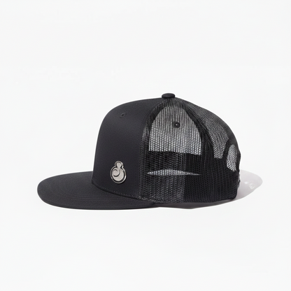 A juice clothing black trucker-style snapback cap with a structured flat brim, black mesh back panels, and a small silver circular logo on the front panel, shown against a plain white background.