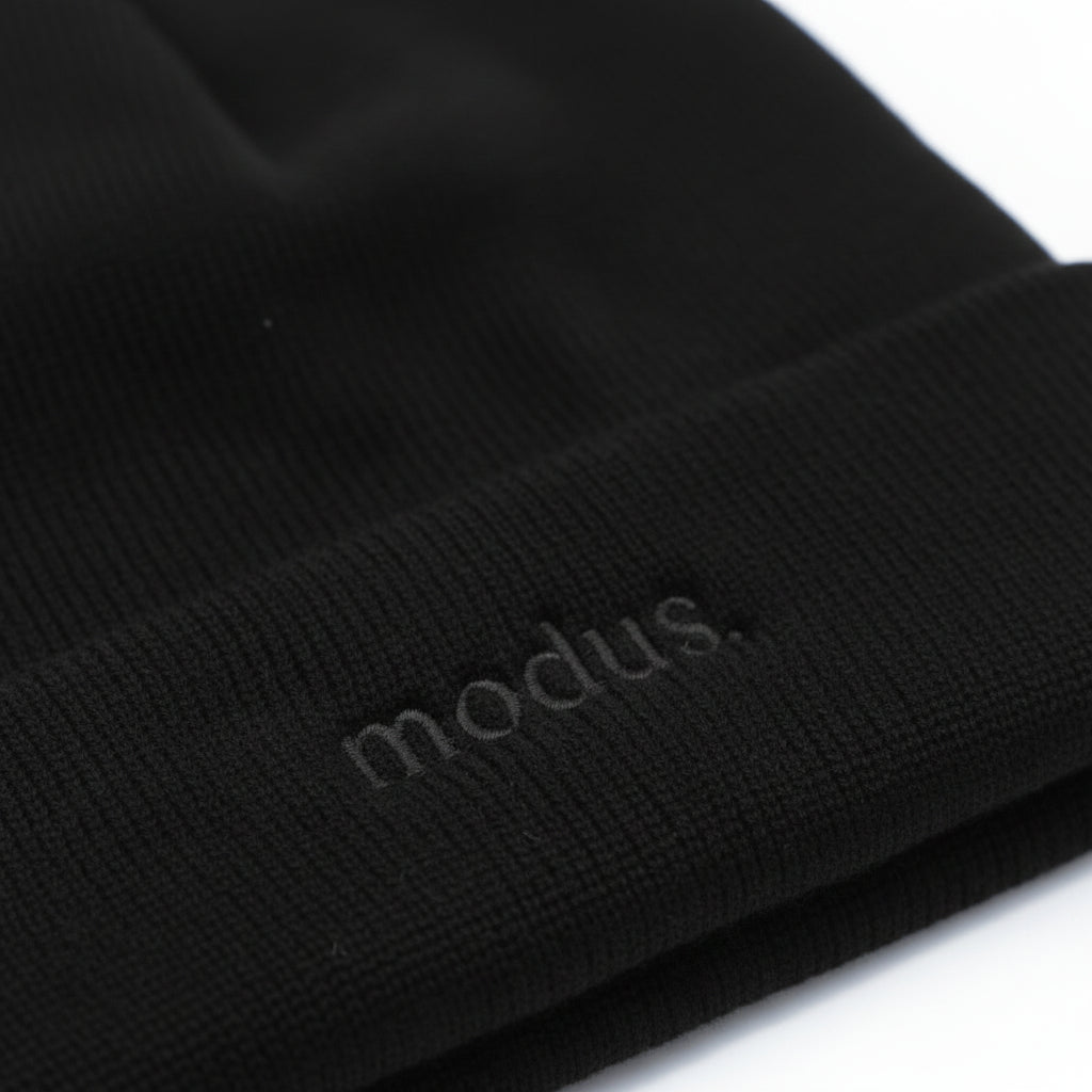 Close-up of a black ribbed knit beanie with the word 'modus.' subtly debossed on the cuff, shown against a white background.
