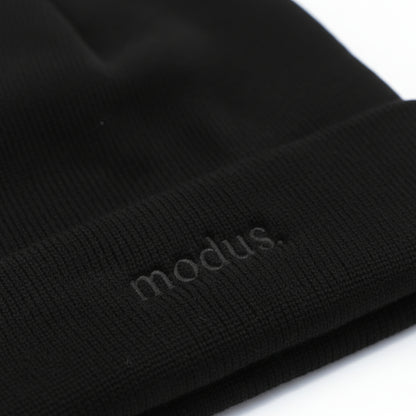 Close-up of a black ribbed knit beanie with the word 'modus.' subtly debossed on the cuff, shown against a white background.