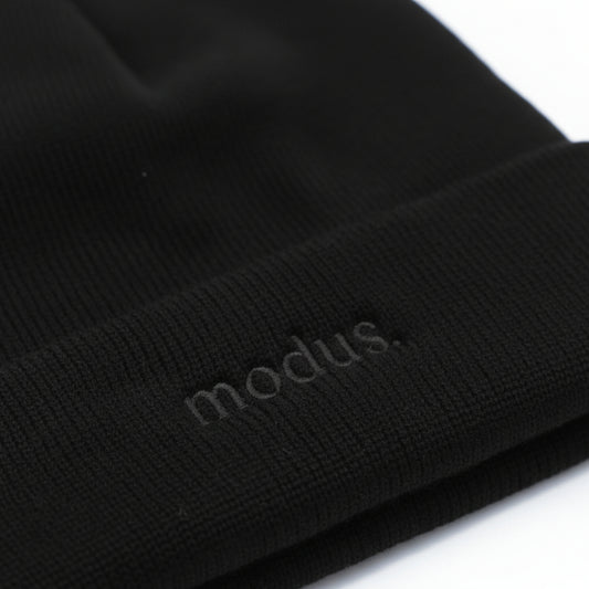 Close-up of a black ribbed knit beanie with the word 'modus.' subtly debossed on the cuff, shown against a white background.